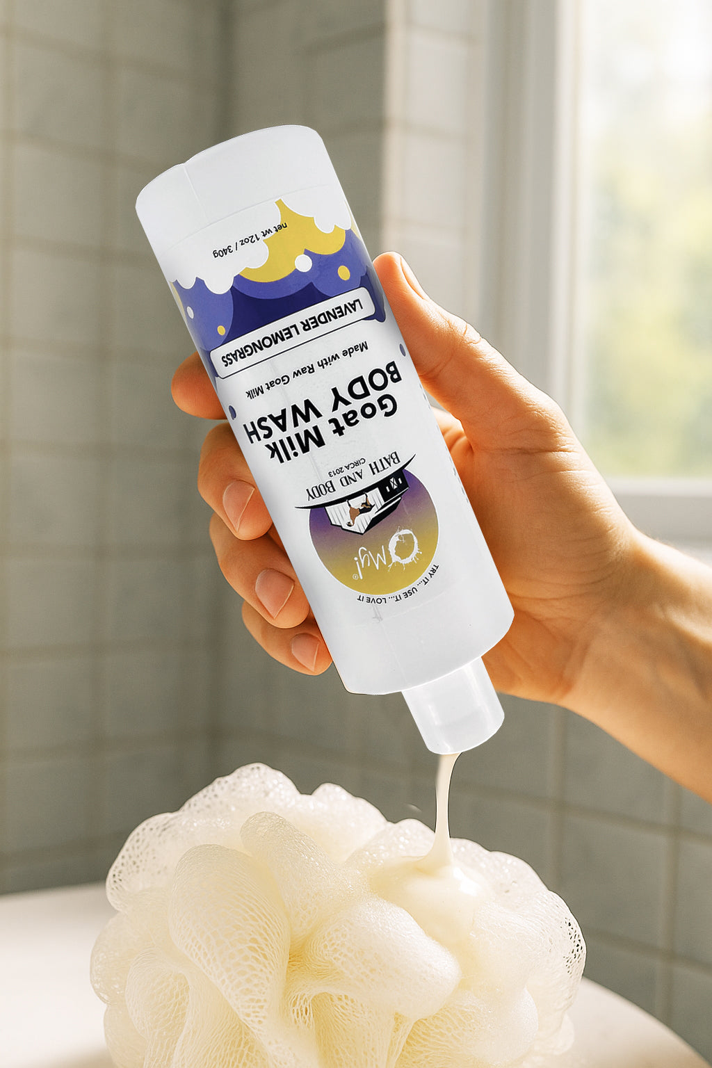 Hand holding a bottle of O My! Goat Milk Body Wash, Lavender Lemongrass essential oil style, with a soap lather on a white background