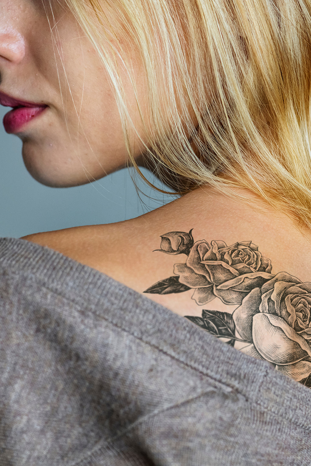 Close-up of a woman's neck with a detailed tattoo of roses on her shoulder against a neutral background.