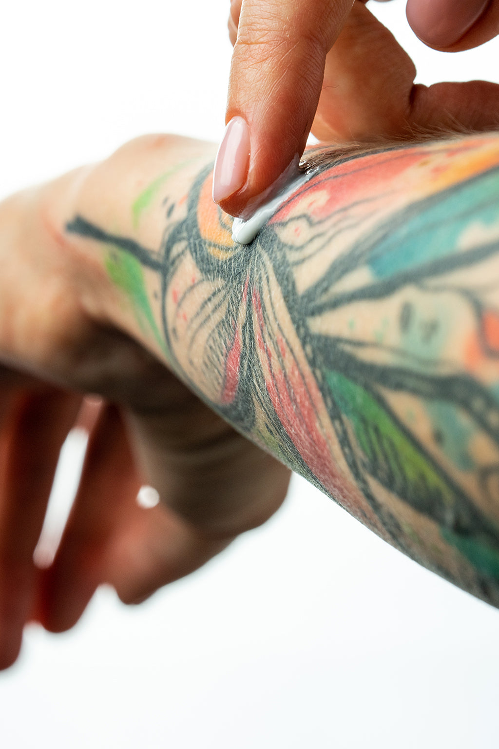 Person applying a white cream or lotion to a colorful tattooed arm on a white background
