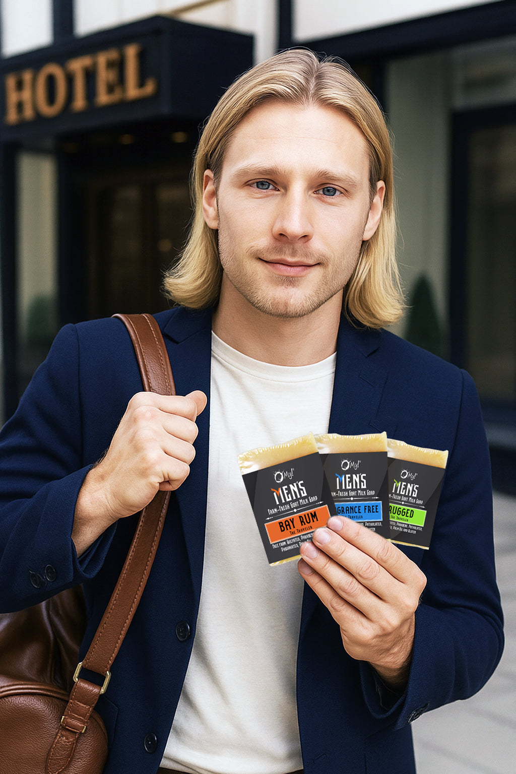Guy holding 3 styles of O My! Men's goat milk traveler soaps in front of a hotel.