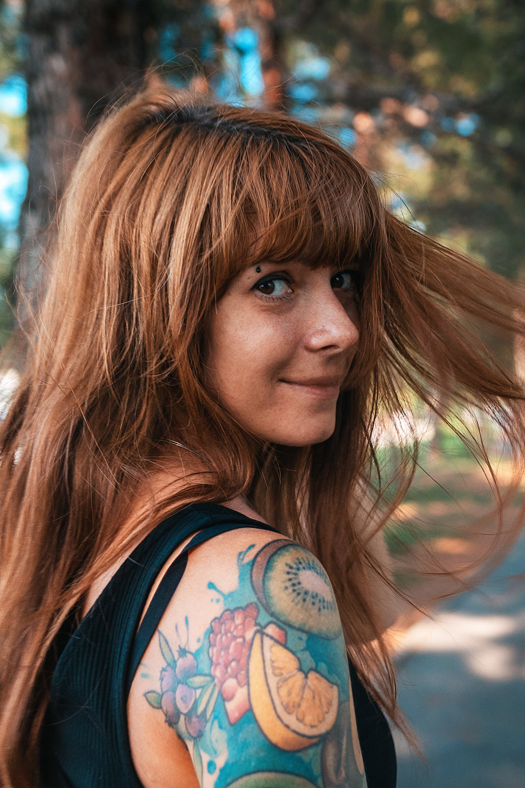 Woman with red hair and  tattoos on her arm standing outdoors with trees in the background