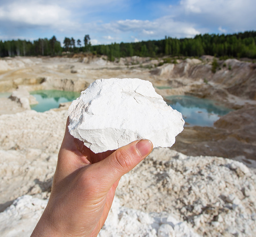 O My! Hand holding kaolin clay with a natural landscape featuring geothermal pools and greenery in the background.