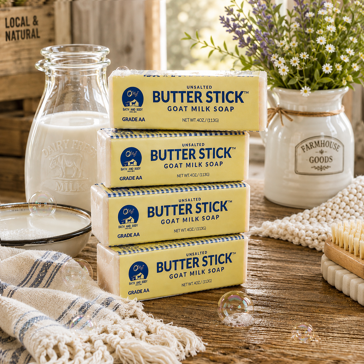 Four stacked bars of O My! Butter Stick Goat Milk Soap on a rustic wooden table, surrounded by a glass milk bottle, a white floral jar, a striped cloth, and soap-dish soap bars.