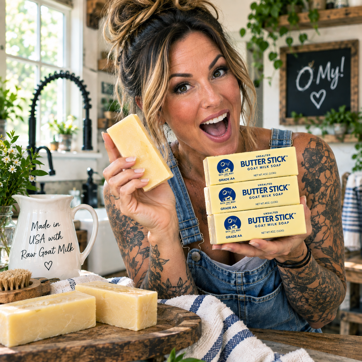 A woman in denim overalls holds three O My! Butter Stick goat milk soap bars in one hand and a large bar of soap in the other, standing in a bright kitchen with more soap bars on the table.