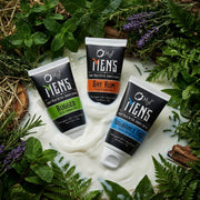 Three tubes of O My! Men's grooming after-shave lotion surrounded by greenery on a white, milky surface.