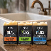 Three bars of O My! Men's goat milk soap with different scents on a wooden countertop by a sink.