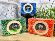 A shaving soap gift box with three bars of soap, each bar has a different color packaging: blue, green, and orange. The bars are presented in front of a gray background with a decorative plant to the side.