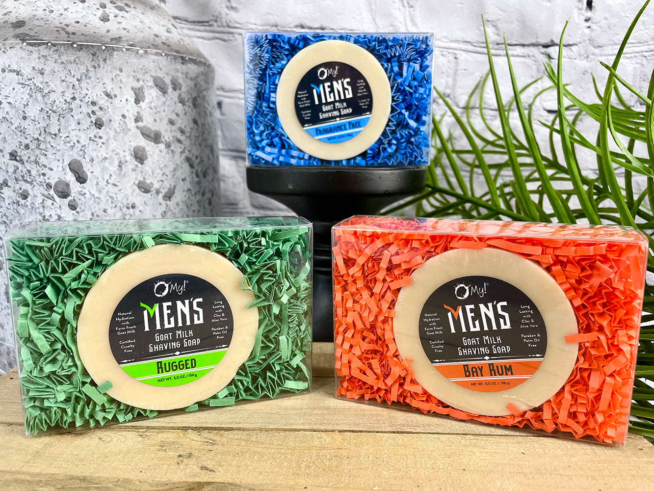 A shaving soap gift box with three bars of soap, each bar has a different color packaging: blue, green, and orange. The bars are presented in front of a gray background with a decorative plant to the side.